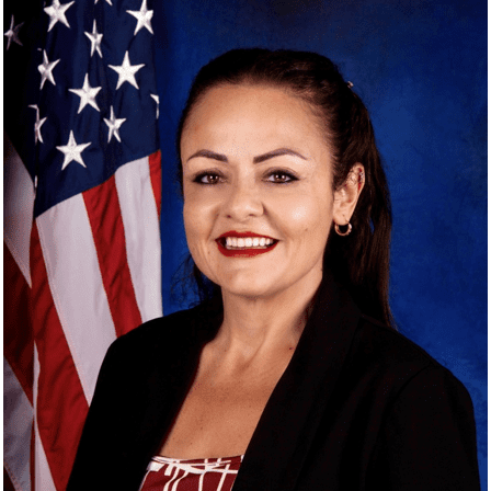 A woman with long dark hair and a bright smile is wearing a black blazer, standing in front of an American flag with a blue background.
