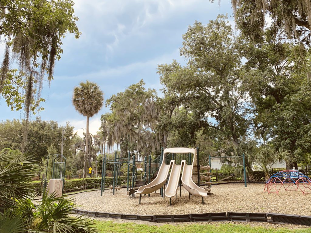 A playground featuring two slides surrounded by trees and a sandy area, with a cloudy sky in the background.