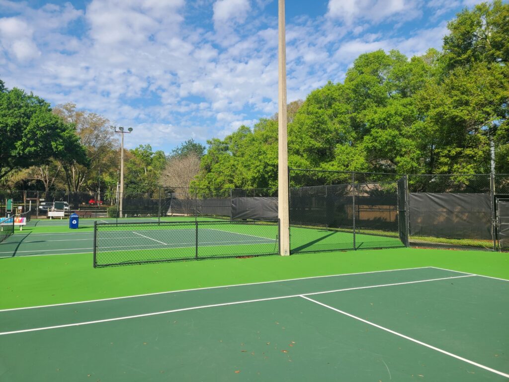 A view of a tennis court with green surfaces, surrounded by trees and a blue sky with scattered clouds.
