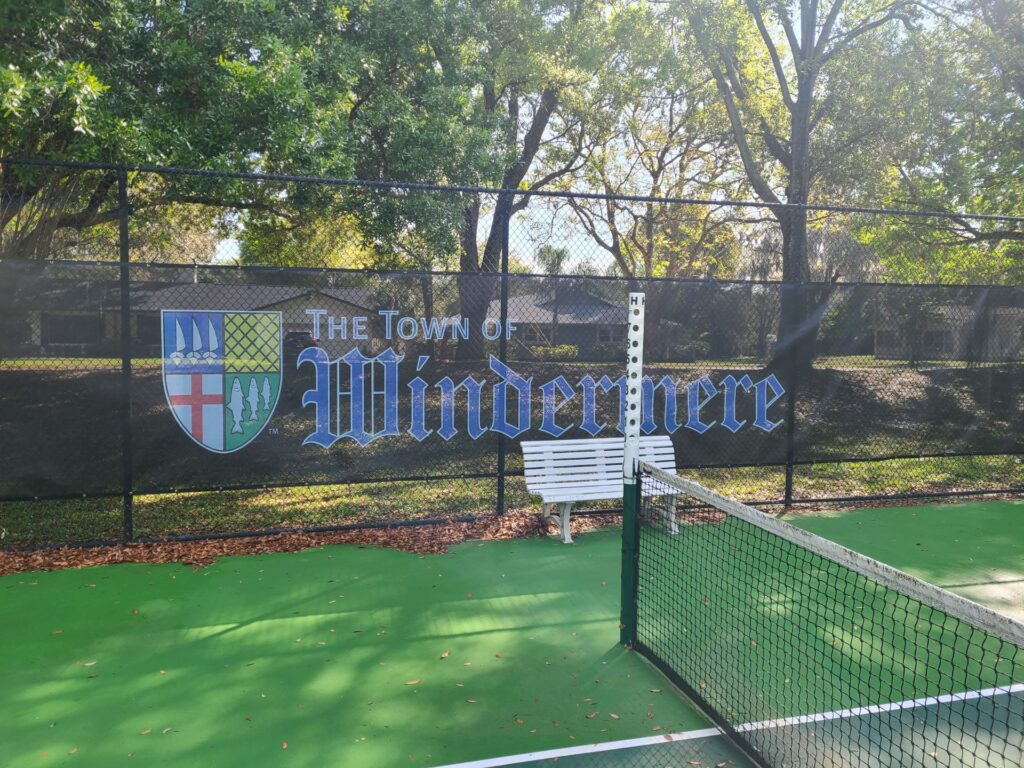 A tennis court featuring a backdrop with the logo and name of 'The Town of Windermere' displayed prominently on a fence.