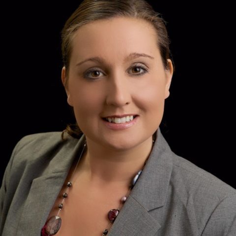 A woman with brown hair, wearing a gray blazer and a necklace, smiles at the camera against a black background.