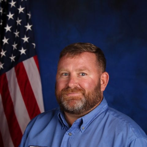 A man with a beard wearing a blue shirt poses in front of an American flag, set against a dark blue background.