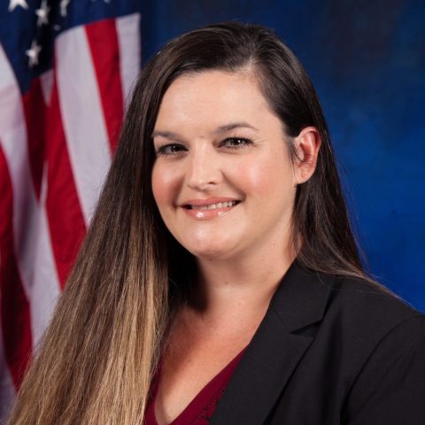 A professional portrait of a woman with long dark hair, wearing a blazer, smiling in front of an American flag backdrop.