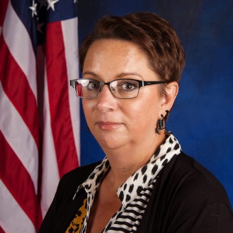 A woman with short brown hair and glasses poses in front of a blue backdrop with American flags.