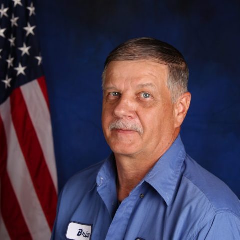A man with short gray hair and a mustache is wearing a blue work shirt with name tags, standing in front of an American flag.