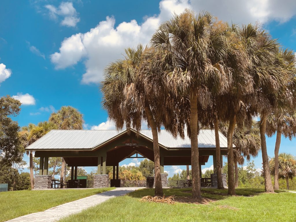 A pavilion surrounded by palm trees, set against a blue sky with fluffy white clouds.