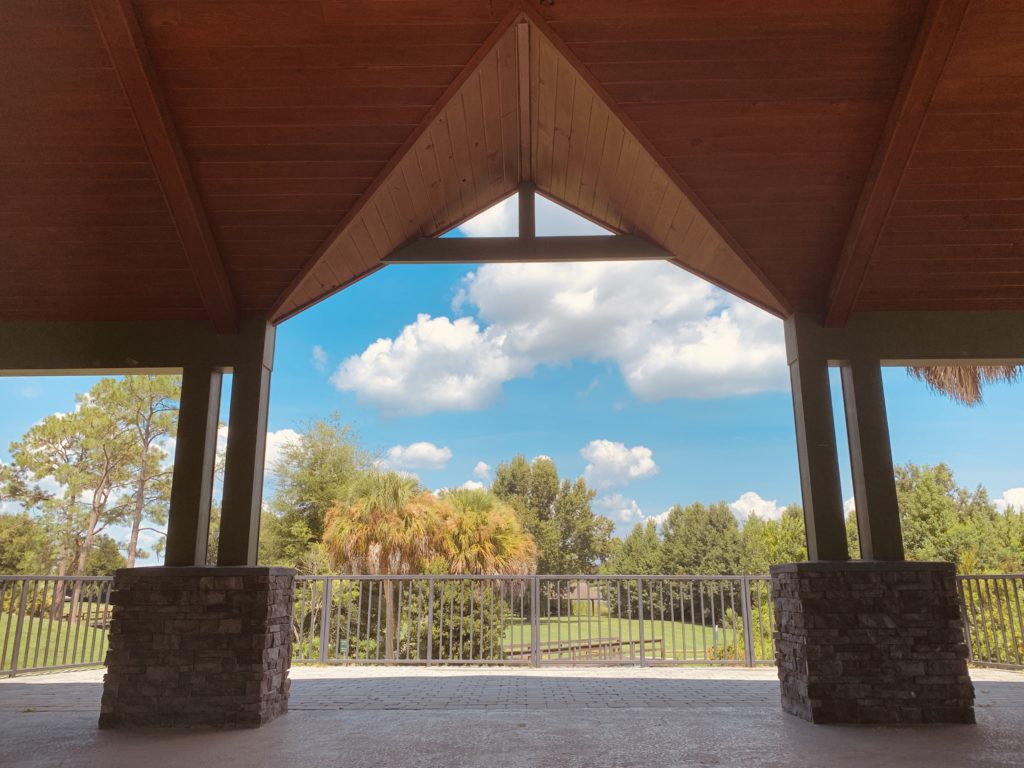 A view from inside a pavilion, showcasing a clear blue sky with fluffy white clouds and lush greenery in the background.
