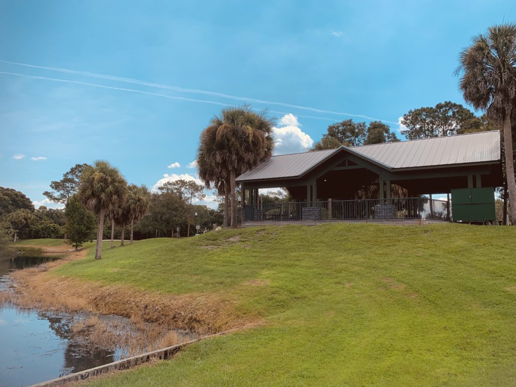 A pavilion with a metal roof is situated on a grassy hill beside a calm waterway, surrounded by palm trees under a clear blue sky.