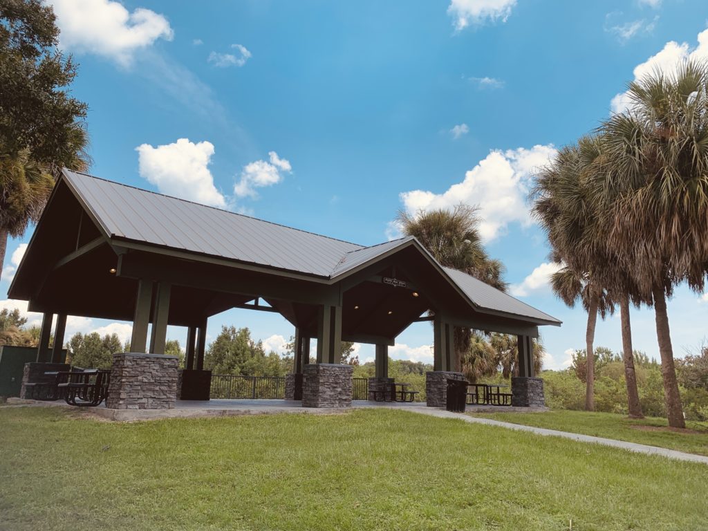 A pavilion with a sloped roof and stone columns, surrounded by green grass and palm trees under a blue sky with fluffy clouds.