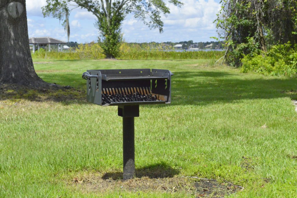 A black charcoal grill mounted on a post, situated on a grassy area with trees and a body of water in the background under a partly cloudy sky.