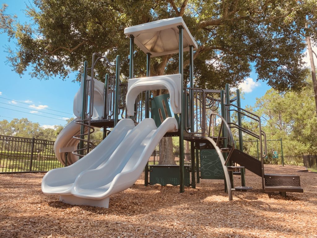 A playground structure featuring two silver slides, surrounded by wood chips and shaded by trees, with a fenced area in the background.