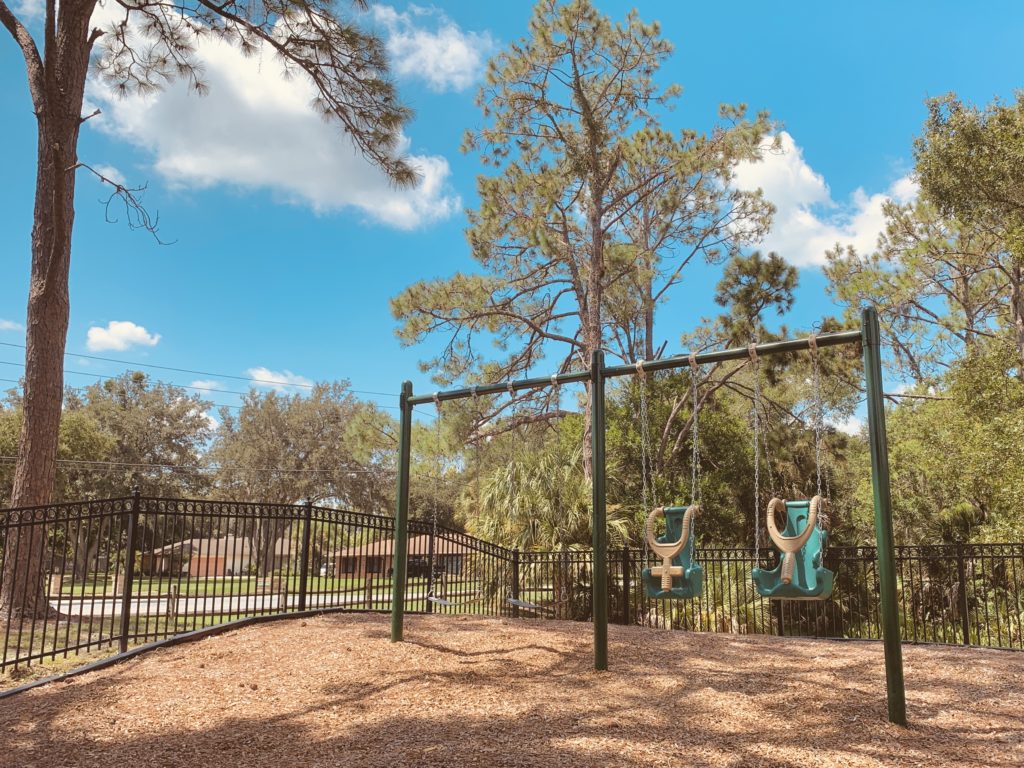 A playground featuring two swings set against a backdrop of trees and a clear blue sky, with a fenced area and a ground covered in wood chips.