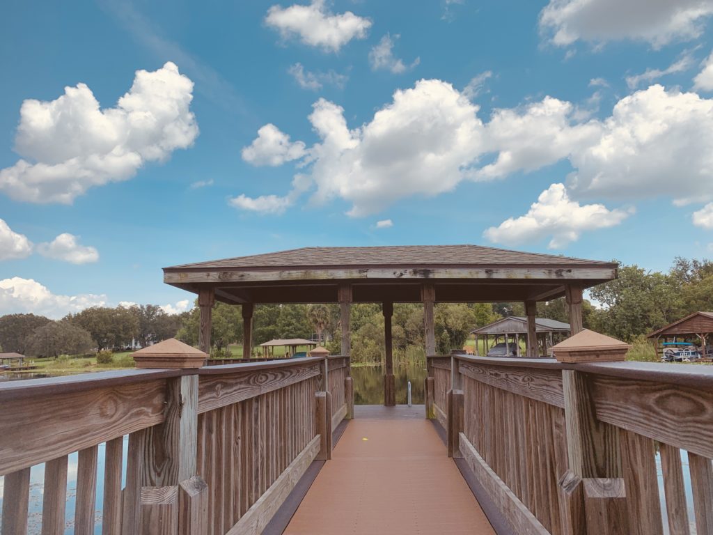 A wooden dock leads to a gazebo overlooking a serene body of water, with fluffy white clouds scattered across a bright blue sky.