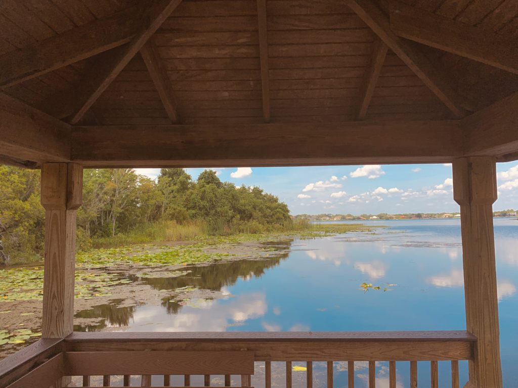 A serene view from a wooden gazebo overlooking a calm lake surrounded by greenery and blue skies with fluffy clouds.