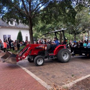 A red tractor with a front loader is parked on a brick path, surrounded by a crowd of people and trees, with a building visible in the background.