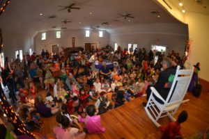 A large group of children sitting on the floor in a hall, attentively watching a person seated in a rocking chair at the front, with decorations and lights around the room.