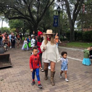 A woman in a wide-brimmed hat walks with two children, one dressed as Spider-Man, in a festive outdoor setting filled with people in costumes.