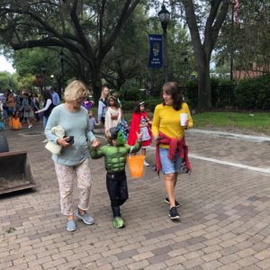 Two women walk along a brick pathway with a child dressed in a green superhero costume, while other children in costumes are seen in the background.