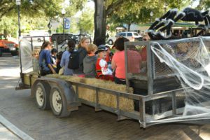A group of children and adults dressed in costumes are sitting on a hay-filled trailer decorated with spider webs, parked under a tree.