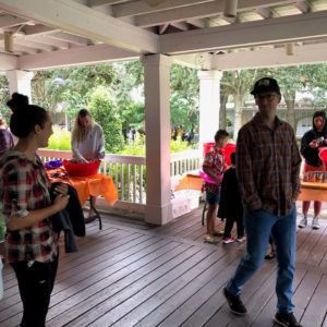 A group of people is gathered under a covered porch, with some individuals interacting and others preparing activities at tables decorated with orange tablecloths.