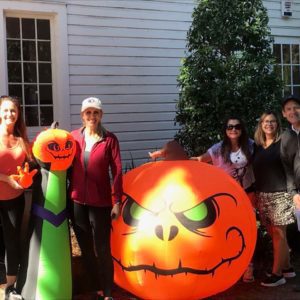 A group of five people poses outdoors next to large inflatable Halloween decorations, including a grinning pumpkin and a character with green and black attire.