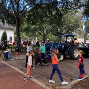 A lively outdoor scene featuring children walking along a path, with a blue tractor parked nearby and a crowd of people in the background enjoying a community event under the trees.