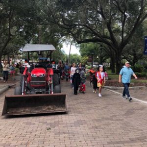 A red tractor is parked on a brick pathway, with people in costumes walking by under trees in a festive atmosphere.