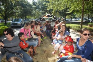 A group of adults and children sitting on a hayride, surrounded by trees and parked cars, enjoying a sunny day.