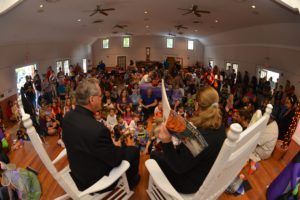 A wide-angle view of a crowded indoor space where two adults sit in rocking chairs facing a large group of children gathered for an event.