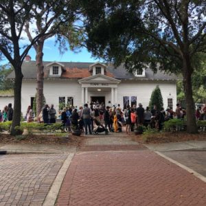 A large crowd gathers outside a white building surrounded by trees, with people dressed in various outfits, suggesting a social event or gathering.