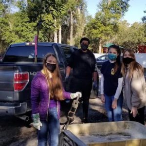 A group of four people wearing masks stands beside a truck, with one person holding a tree sapling, while a wheelbarrow is in front of them.
