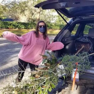 A person wearing a pink sweatshirt and a mask stands beside a car with branches and greenery in the trunk, smiling and posing playfully.