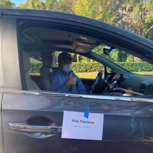 A person wearing a mask gives a thumbs-up from the driver's seat of a car, which has a sign reading 'Pink Table' attached to the window.