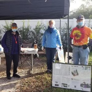 Three individuals wearing masks stand outdoors under a canopy, with a table and a poster displayed in front of them.