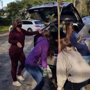 A group of four people are gathered around the open trunk of a car, assisting a woman in unloading a small tree or plant, while wearing masks and gloves.