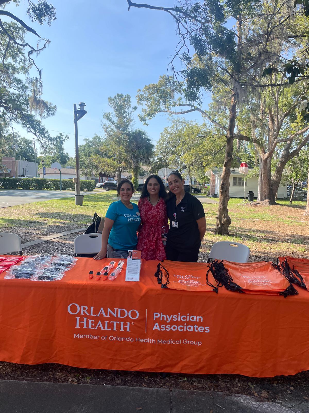 Three women stand behind an orange Orlando Health table with health-related items outdoors in a park.