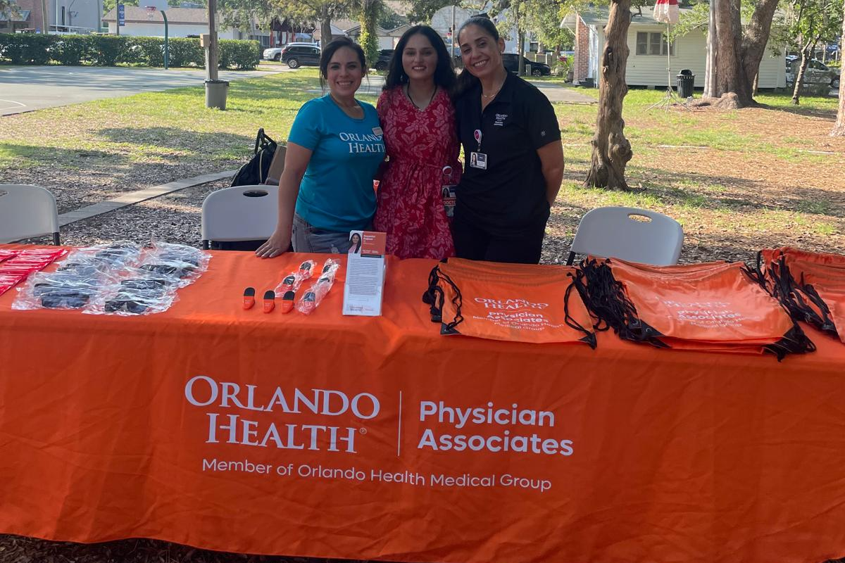 Three women stand behind an orange Orlando Health table with health-related items outdoors in a park.