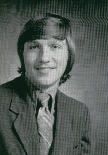 A black and white portrait of a young man with long hair, wearing a suit and tie, smiling at the camera.