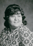 A black and white portrait of a woman with curly hair, wearing a patterned blouse, smiling at the camera.