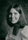 A black and white portrait of a young woman with long, dark hair, smiling at the camera, wearing a light-colored dress with lace details.