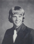 A black and white portrait of a young man with long, wavy hair, wearing a suit and tie, smiling at the camera.