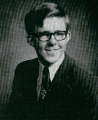 A black and white portrait of a young man with glasses, wearing a suit and tie, smiling at the camera.
