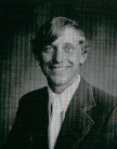 A black and white portrait of a young man with a smile, wearing a suit and tie, set against a textured background.