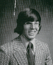A man with long hair and glasses smiles while wearing a patterned suit and tie against a plain background.