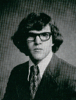 A black and white portrait of a young man with curly hair and glasses, wearing a suit and tie, looking directly at the camera.