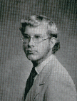 A black and white portrait of a man with glasses and light-colored hair, wearing a suit and tie, posed in profile against a plain background.