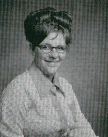 A black and white portrait of a woman with glasses and a large hairstyle, wearing a light-colored blouse and smiling at the camera.