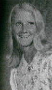 A black and white portrait of a young woman with long, light-colored hair, smiling and wearing a collared blouse with floral patterns.