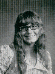 A black and white portrait of a woman with long hair and glasses, smiling at the camera, wearing a patterned blouse.