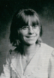 A black and white portrait of a young woman with glasses and shoulder-length hair, smiling at the camera.